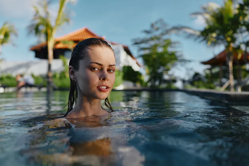 Young woman with wet hair enjoys swimming in outdoor pool near tropical plants and sunny sky, relaxed lifestyle moment captured on a warm day.