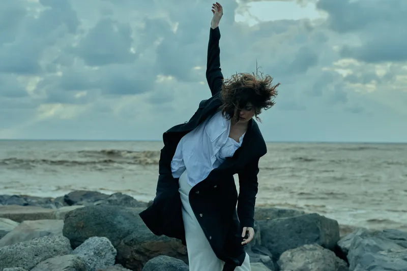 Fashionable figure moves along a rocky shoreline, wearing a long coat and white dress, wind lifting fabric as waves crash, creating a moody cinematic coastal scene with dramatic motion