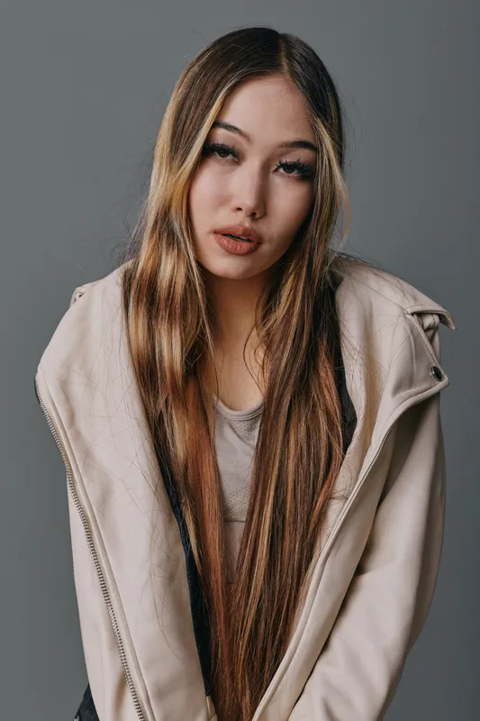 Young woman with long straight hair wearing beige jacket and casual shirt posing with serious expression against gray background in studio portrait.
