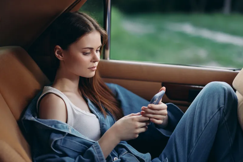 A young woman in casual denim attire relaxing in a car, using her smartphone, expressing calmness in a serene outdoor setting.
