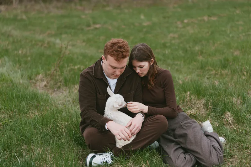 Caring couple outdoors, sitting on grass, softly holding a white goat kid, intimate moment, warm natural light, relaxed posture, gentle interaction, pastoral setting, earthy tones, peaceful