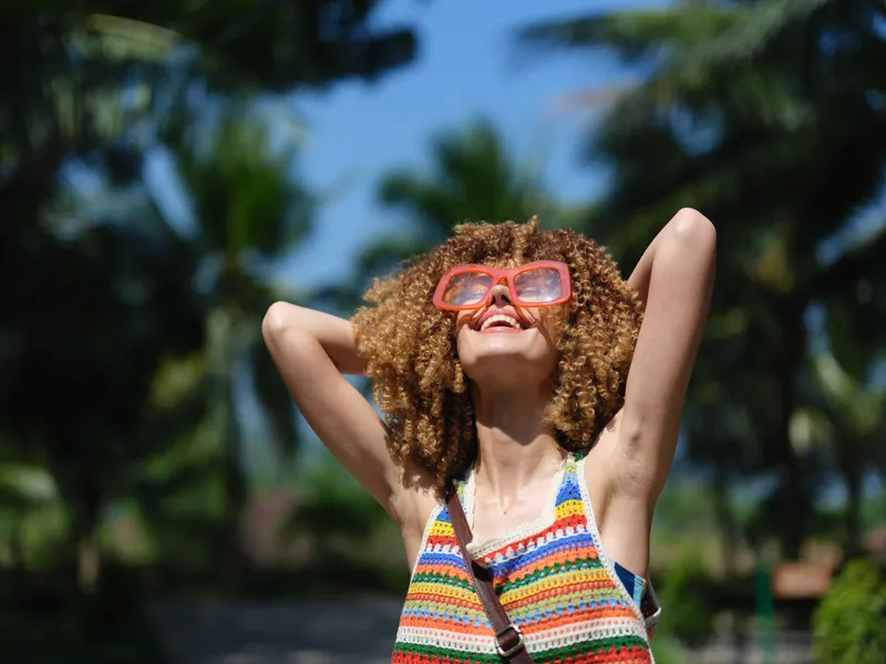 Smiling woman wearing sunglasses enjoys sunny day outdoors with vibrant summer dress and curly hair against tropical palm tree background.