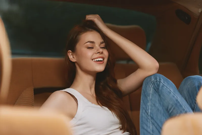 Young woman smiling in a car interior, wearing a casual white tank top and jeans, showcasing a relaxed and happy mood with warm tones highlighting the cozy atmosphere.