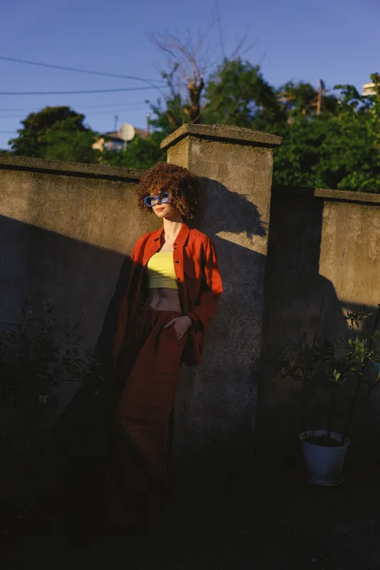 A stylish woman poses outdoors beside a stone wall in a red dress and sunglasses, capturing a casual lifestyle moment with warm sunlight and relaxed confidence