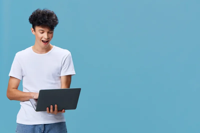 young man using laptop, wearing casual white t-shirt, expressing surprise, curly hair, blue background, indoor technology scene with modern style