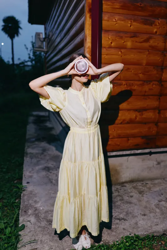 Woman in a pale yellow dress stands beside a wooden building at dusk, holding a camera to her face to create a moody outdoor portrait.