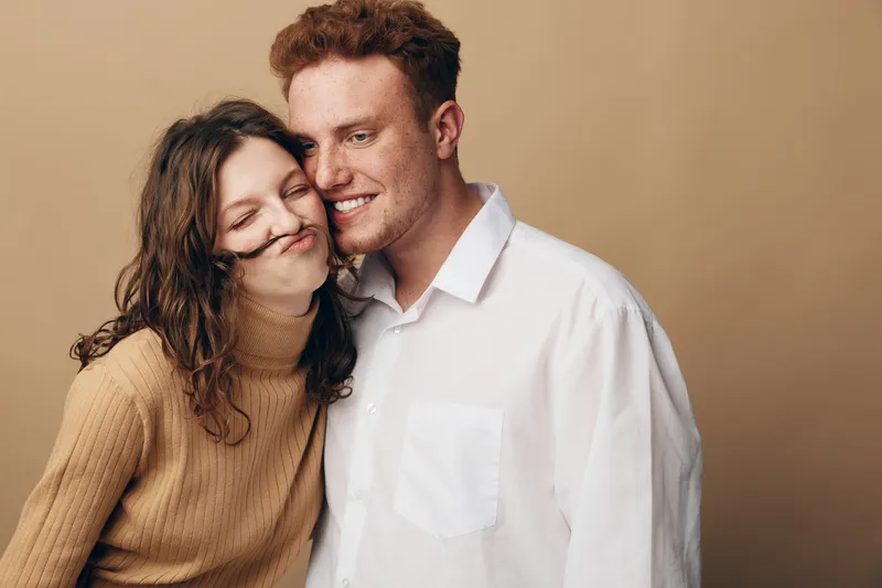 Young couple in casual clothes share a playful moment, woman with curly hair makes a mustache with her hair, man smiles gently. Concept of love, fun, and relationship happiness.