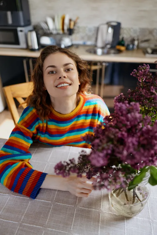 A young woman with curly hair sits at a bright kitchen table in a striped colorful sweater, smiling toward the camera beside a vibrant purple bouquet and fresh flowers.