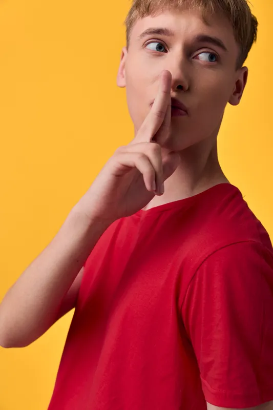 Shushing gesture by a young male in a red shirt against a bright yellow background, conveying silence, secrecy, and quiet communication in a minimal scene