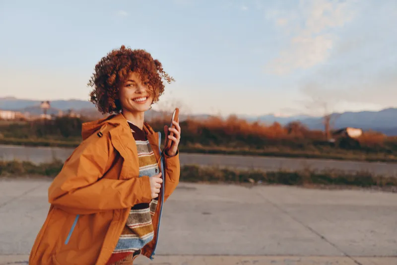 Woman lifestyle outdoors, curly hair, orange jacket, smiling at sunset on a road, holding smartphone for selfie, casual travel portrait, soft light copy space