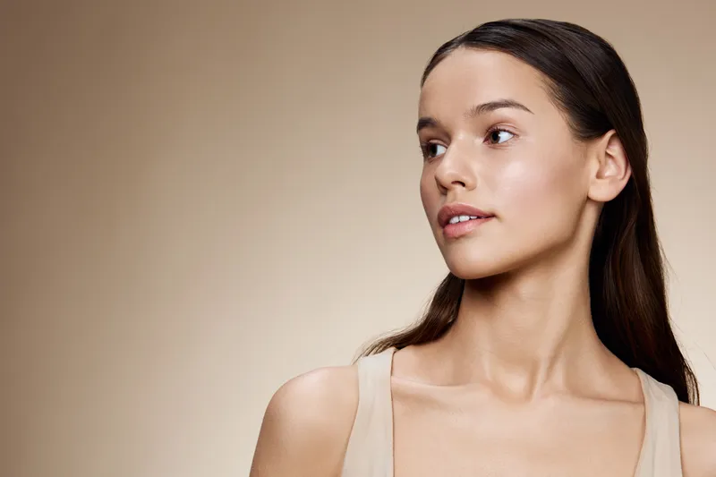 Elegant portrait of a young woman with long brown hair and natural makeup, showcasing beauty and confidence against a soft beige background