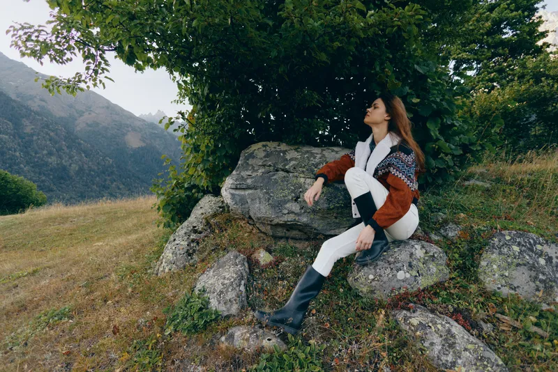 Woman sitting on rocks under a tree in a mountain meadow with mountains in the background, nature scenery and serene landscape, relaxed portrait in sweater and boots enjoying peaceful relaxation.