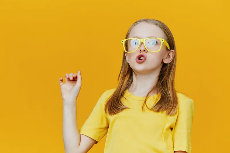 Young girl with glasses expressing excitement in a vibrant yellow outfit against a bold yellow background, symbolizing joy and creativity