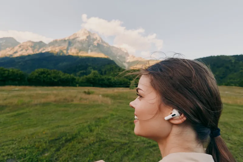 Young woman with brunette hair tied back enjoys music on wireless earbuds while standing in a green field with mountains in the background under a clear sky.