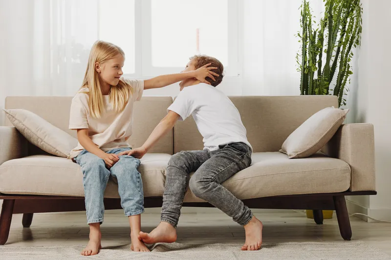 Two children sitting on a beige sofa in a bright living room, girl playfully touching boy’s head. Concept of sibling interaction, childhood, family moments, and playful behavior.