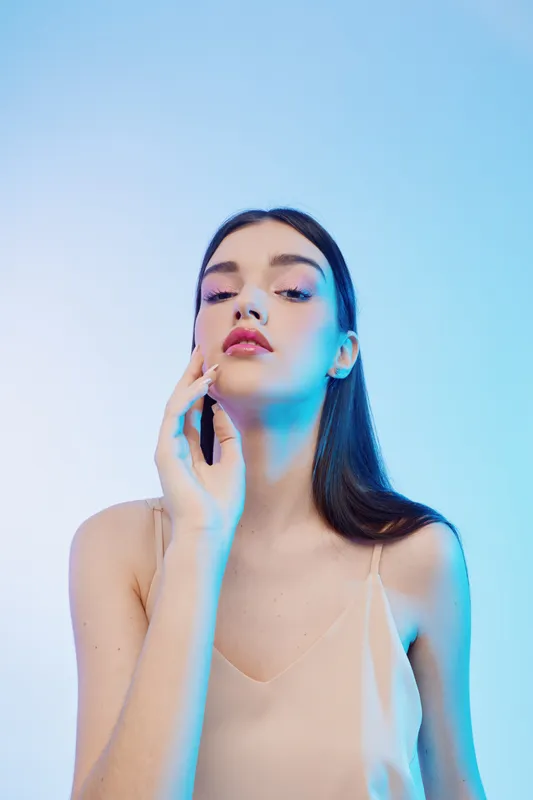 Young woman with long dark hair poses with hand near face against light background. Soft lighting highlights her clear skin and elegant beauty. Concept of woman beauty fashion.