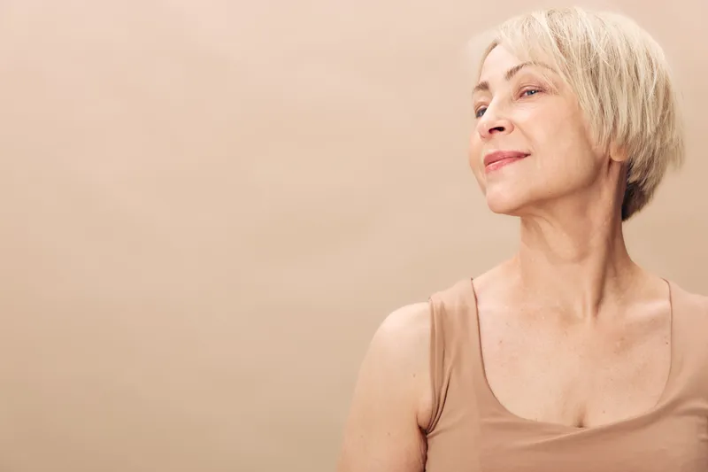 Smiling mature woman with short blonde hair looks away thoughtfully on beige background. Concept of aging beauty, natural skin, confidence, and positive lifestyle for older adults.