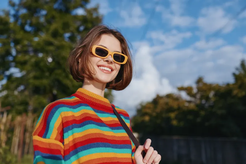 Woman in rainbow sweater and yellow sunglasses smiling outdoors under blue sky, short hair bob, casual outfit, sunny lifestyle portrait in park, joyful travel vibe and confidence.