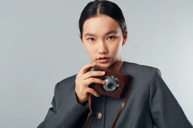 Young woman holding vintage camera close to face with intense look wearing gray blazer against neutral background. Portrait, photography, fashion, retro, technology concept.