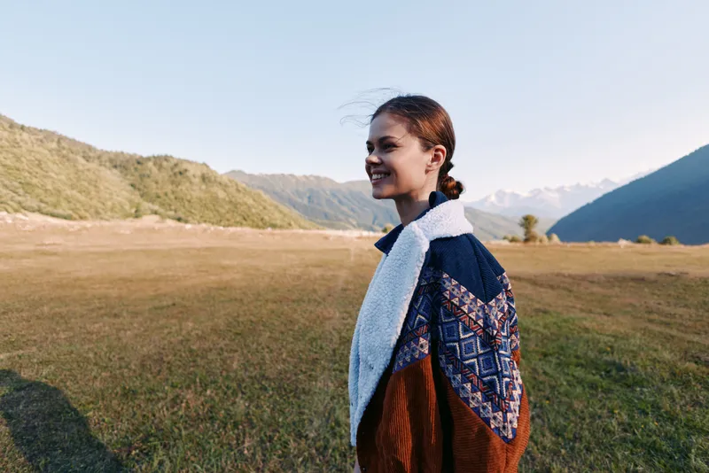 Woman, smile, landscape, mountains, meadow, sweater, outdoors, portrait — Young woman smiling in a cozy sweater standing in a meadow by mountain landscape at golden hour for travel and nature.