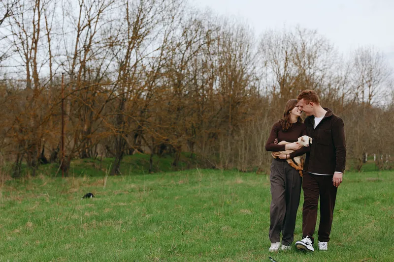 Young couple embraces tenderly in a green field while holding a small baby goat, enjoying a peaceful moment outdoors with bare trees in the background during early spring.