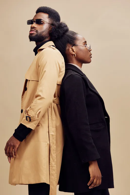 Stylish couple in modern attire with sunglasses standing back to back, showcasing a blend of fashion and confidence in an elegant neutral background