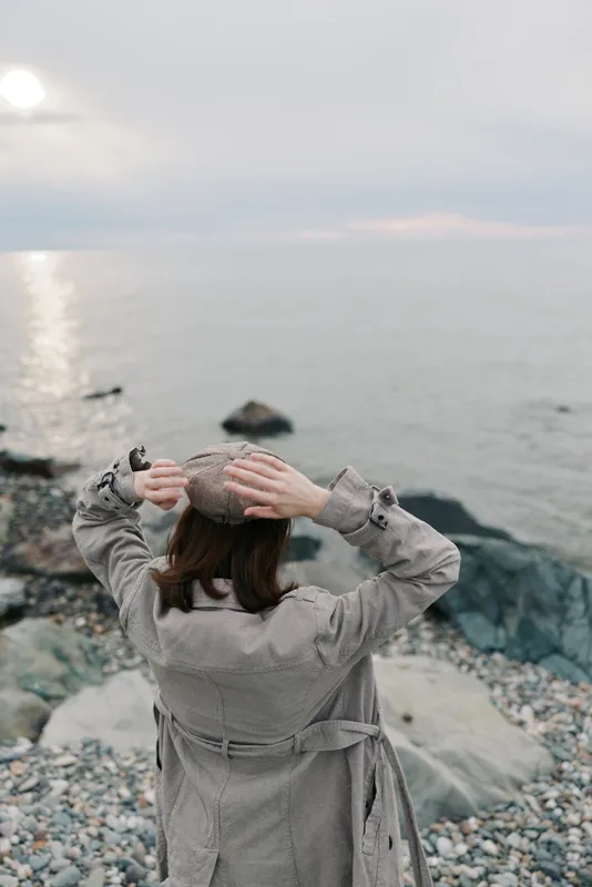 A woman in a gray coat and hat stands on a rocky shore facing the sea, holding her hat with both hands. The calm water and cloudy sky create a peaceful coastal scene.
