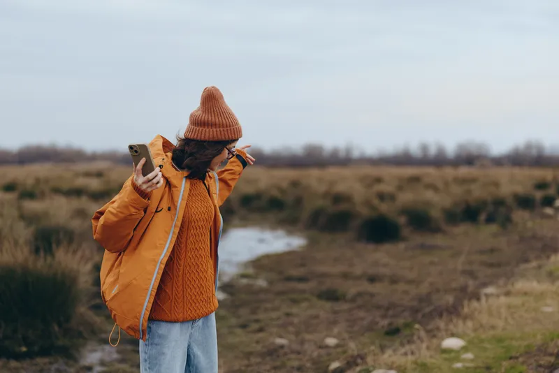 Woman lifestyle outdoors in rural field, wearing winter coat and knitted hat, walking and looking down, calm nature, cloudy sky, natural colors, path and shrubs, soft breeze, cool