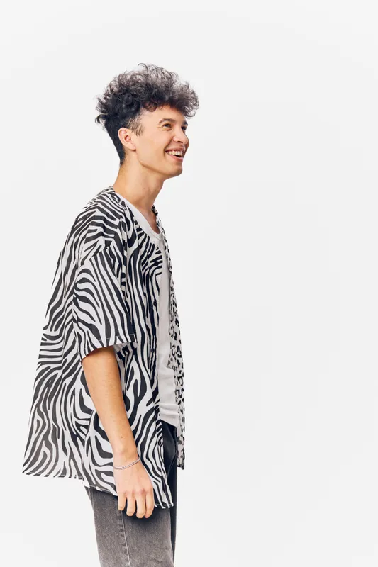 Smiling man wearing zebra print shirt and jeans stands against white background, showing joyful expression and casual modern style with curly hair and bracelet.