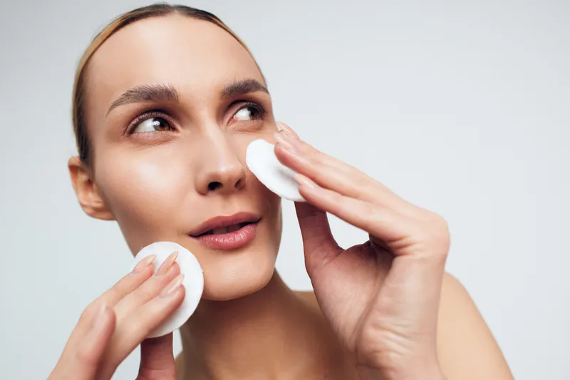 A woman applies a cotton pad to her face in a close-up studio shot. Clear skin, minimal makeup, soft lighting conveys skincare, wellness, and self-care with a focused beauty mood.