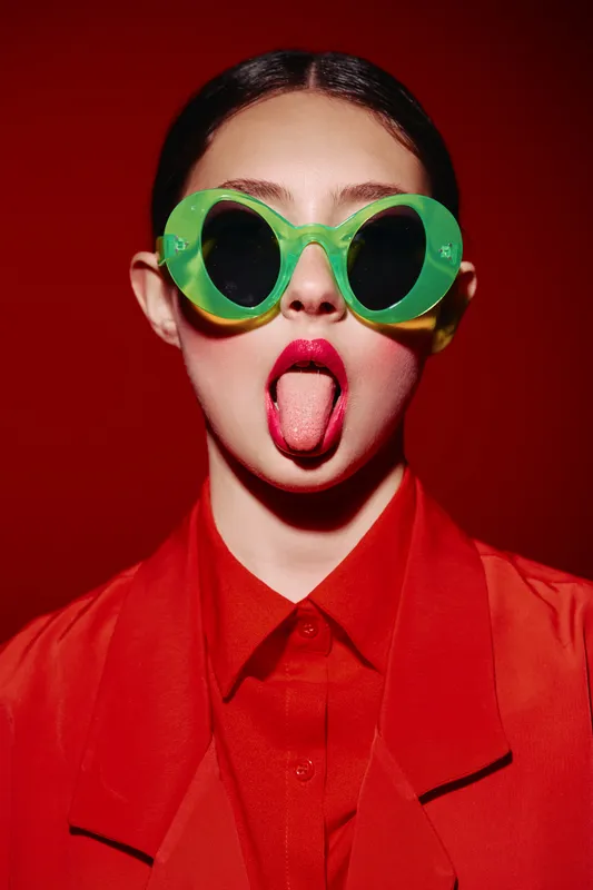 Young woman wearing oversized green sunglasses and red lipstick sticks out tongue on a red background. Studio fashion portrait conveying playful and bold beauty creativity.