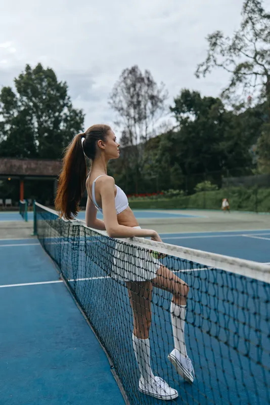 Young athletic woman in activewear standing by a tennis net, showcasing confidence and dedication on a beautiful outdoor court background