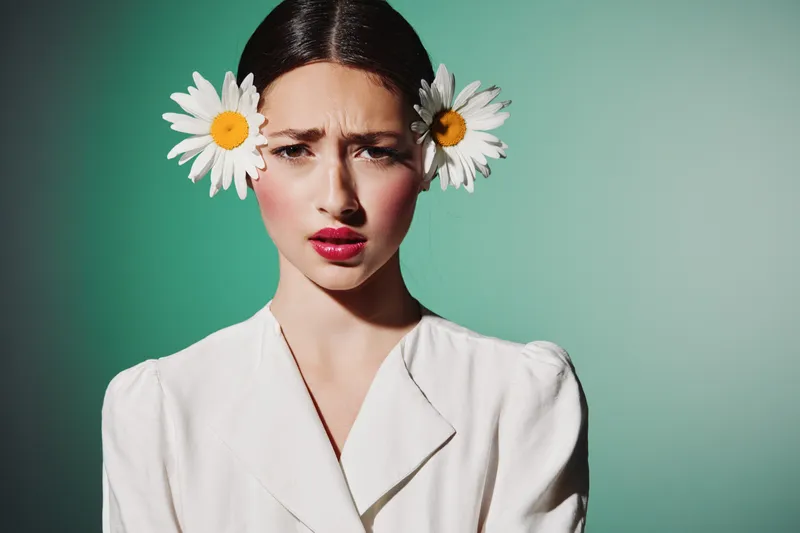 Young woman in white with daisies in hair looks serious studio portrait on green background. Bright makeup and floral fashion creative concept for beauty style photography.