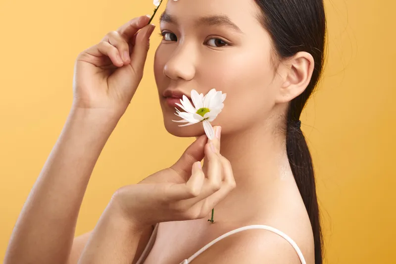 asian woman with dark hair holds a white daisy near her lips against a warm yellow background, conveying calm beauty and gentle charm in a studio portrait.