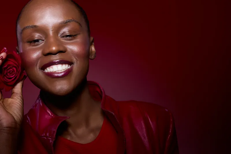 Smiling dark skinned African woman holds red rose near face, wearing red leather jacket and top. Studio portrait on red background. Concept of beauty, happiness, and natural skin glow.