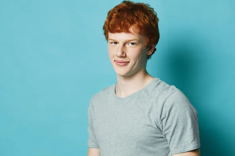 Young man with red hair and light skin wearing grey t-shirt against blue background looking at camera with slight smile in studio portrait photography.