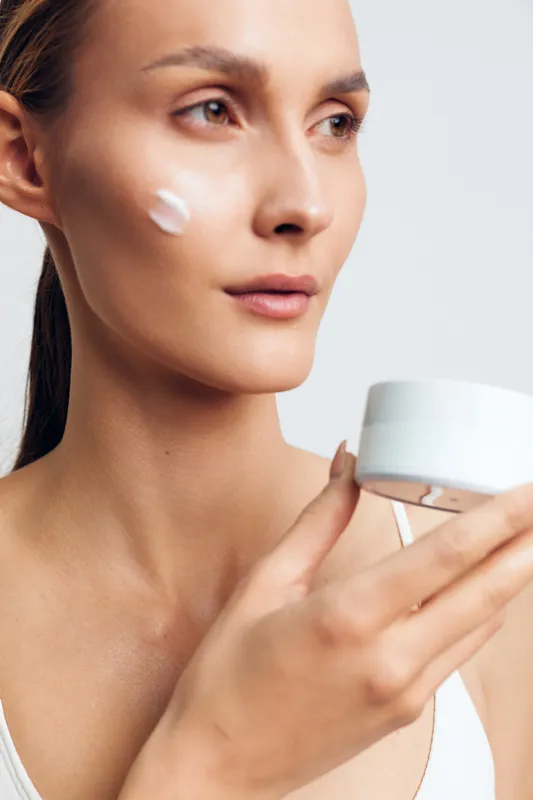 A close-up portrait of a young woman applying moisturizer to her cheek in a bright studio, conveying a focused skincare ritual and a calm, confident mood with clean background and soft light.