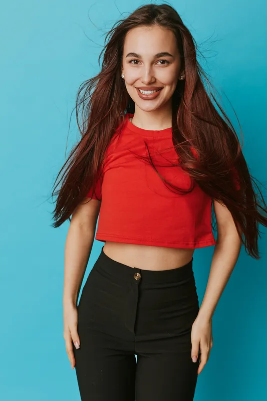 Young woman with long hair wearing red crop top and black pants smiling confidently standing against blue background in studio. Casual fashion, beauty and positive expression portrait.