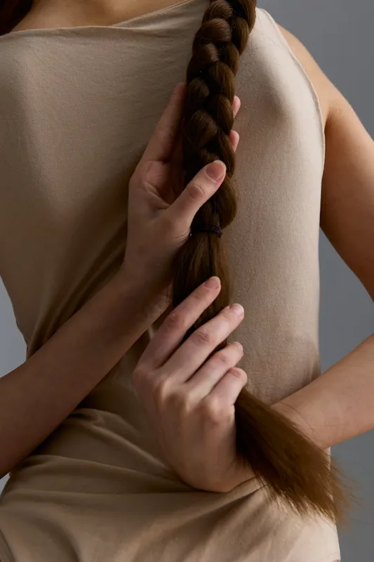 Woman with long braided hair wearing a beige textured sleeveless dress, holding braid with graceful hands against a neutral gray background, natural and calm mood.