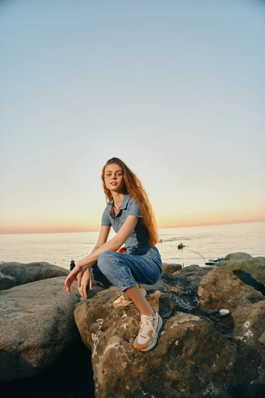 lifestyle woman in modern boho-western denim street style poses on rocks by the sea at sunset with warm film color tones
