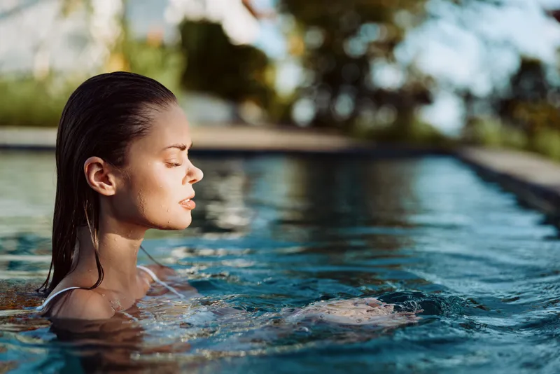Young woman with braided hair relaxes in outdoor pool water with closed eyes enjoying sunny day, calm lifestyle moment, blurred natural background visible.