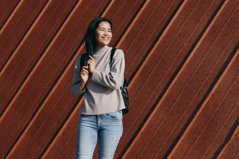 Woman with a backpack, casual denim outfit stands in front of a diagonal wooden wall, smiling, outdoor street style, relaxed fashion vibe for a candid urban moment