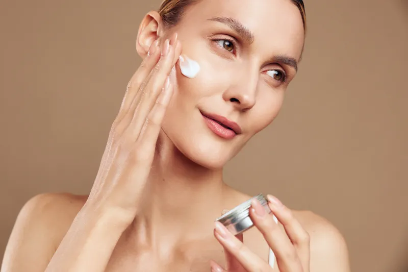 Young woman applies face cream on clean skin with gentle touch, holding jar of moisturizer, shows skincare routine and natural beauty on beige background, closeup portrait