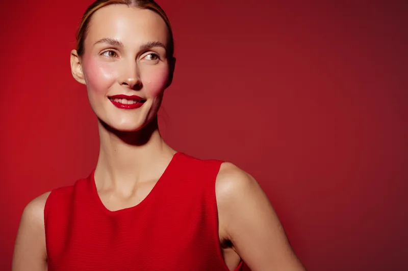 Smiling woman in red sleeveless dress poses against red background. She has bright red lipstick and smooth skin, creating a vibrant and elegant portrait with warm lighting.