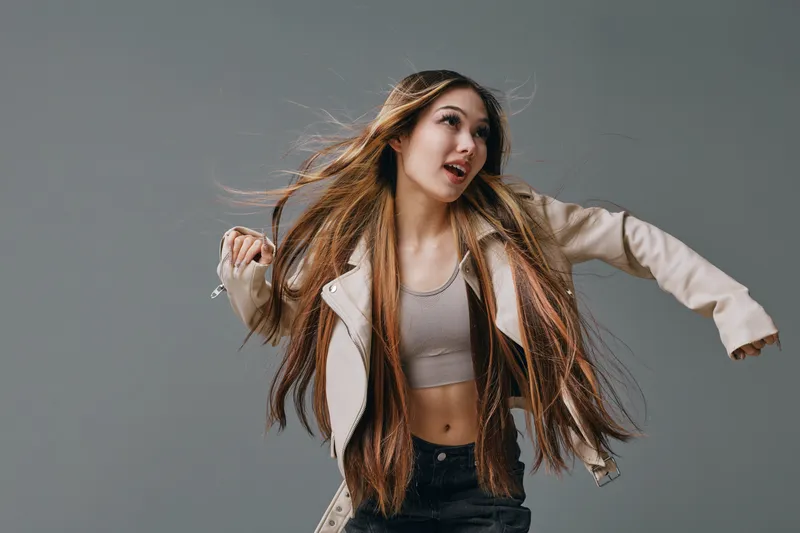 Woman with long hair wearing jacket and crop top dancing on gray studio background with energetic expression and motion captured in mid action.