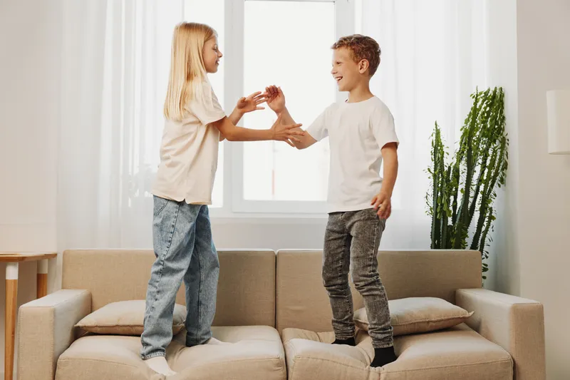 Two happy children standing on couch holding hands smiling in bright living room with plant and window background. Concept of family fun, sibling bond, and playful childhood.