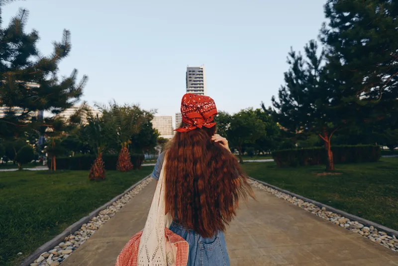 Lifestyle woman in modern boho-western denim street style walks outdoors with long wavy hair and red bandana, surrounded by green trees and urban park setting, film color.