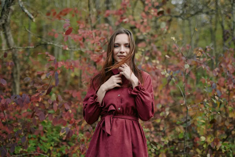 Portrait of a young woman in a burgundy dress among autumn foliage, captured with authentic natural light and a genuine expression that conveys honesty, intimacy and emotional realism.