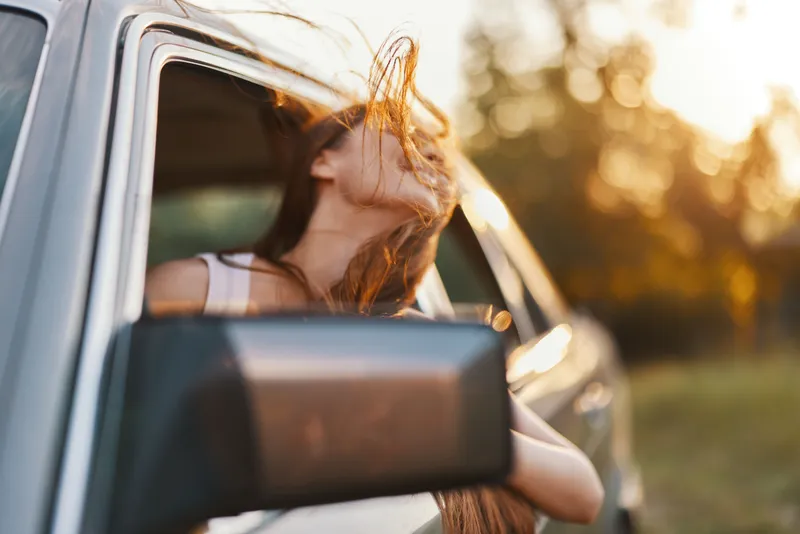 Young woman enjoying freedom while driving a car with wind in her hair, vibrant sunset in background, evoking feelings of adventure and joy.