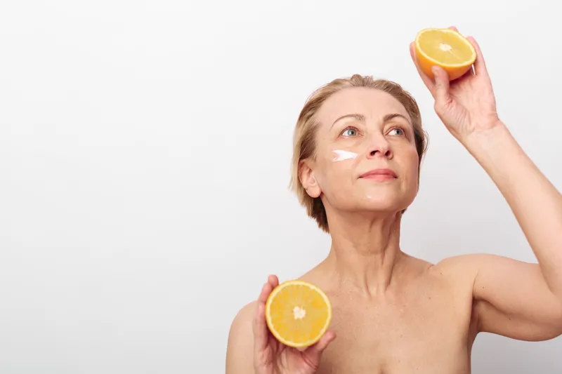 Mature woman with natural skin holds orange halves near face with cream on cheeks. Bright simple background highlights skincare, healthy lifestyle, and natural beauty concepts.